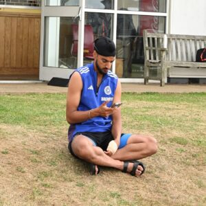Indian fast bowler Arshdeep Singh sits on the grass with a bandaged wrist, looking at his phone.