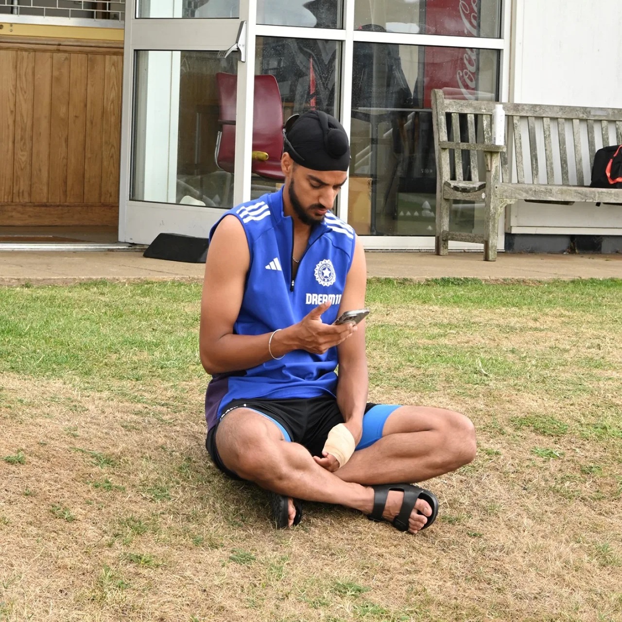 Indian fast bowler Arshdeep Singh sits on the grass with a bandaged wrist, looking at his phone.