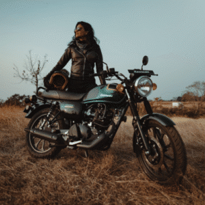 A woman rider standing beside a Kawasaki W175 motorcycle in an open field, wearing a leather jacket and holding a helmet