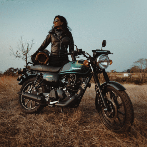 A woman rider standing beside a Kawasaki W175 motorcycle in an open field, wearing a leather jacket and holding a helmet